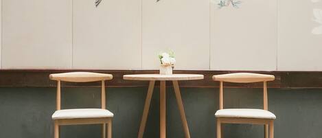Una habitación con dos sillas de madera y una mesa redonda, un florero con flores, y una pared decorada con pájaros y flores pintados.