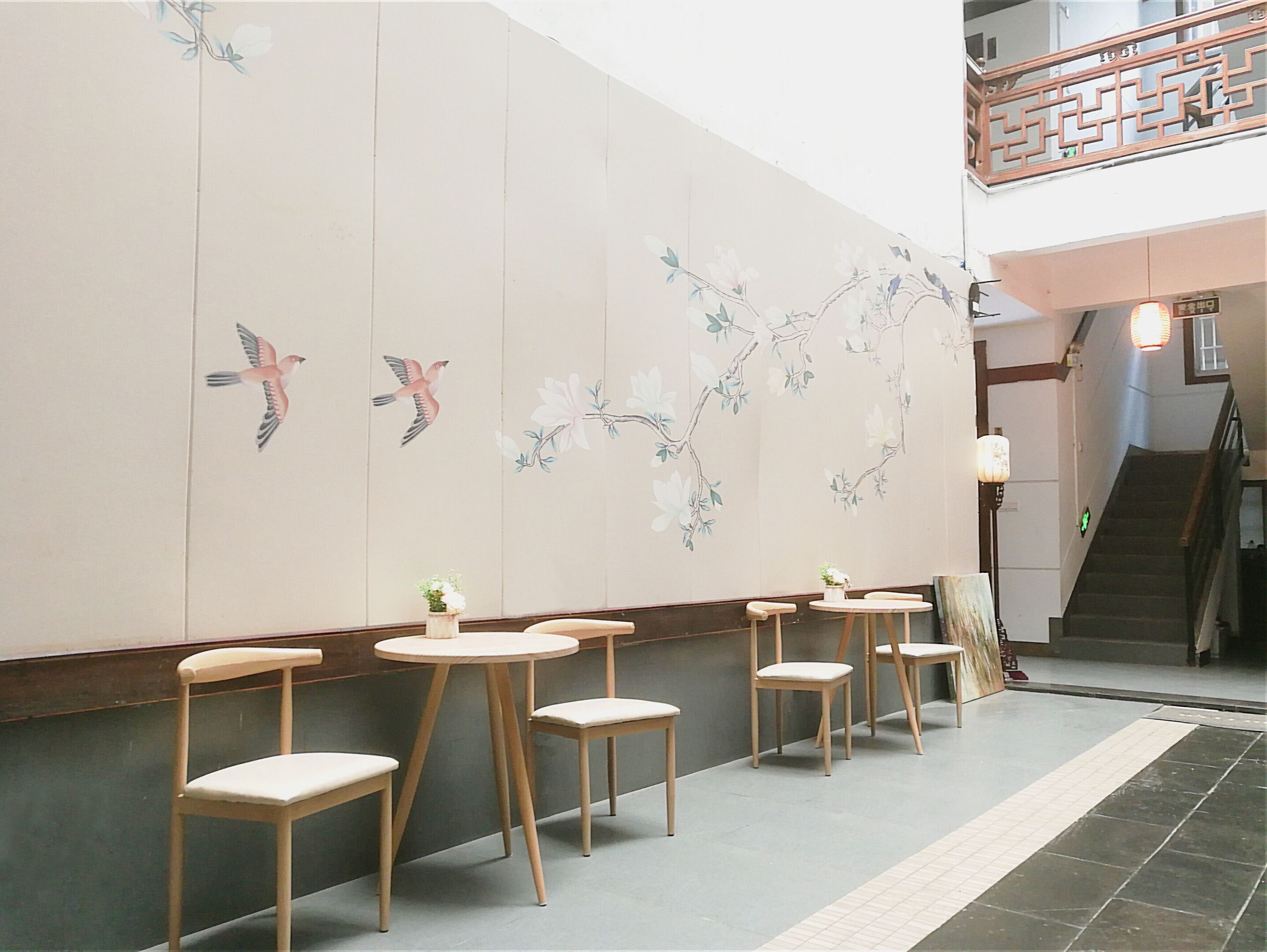 Un restaurante con sillas y mesas de madera, una pared decorativa con murales de pájaros y flores, y una escalera al fondo.