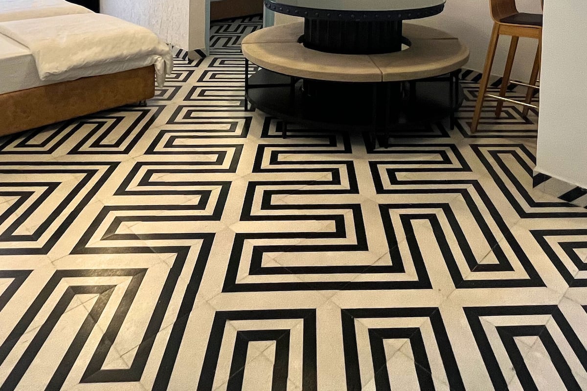 Une pièce dotée d’un sol à motifs géométriques, d’un lit recouvert de draps blancs et d’une table ronde à piétement noir.