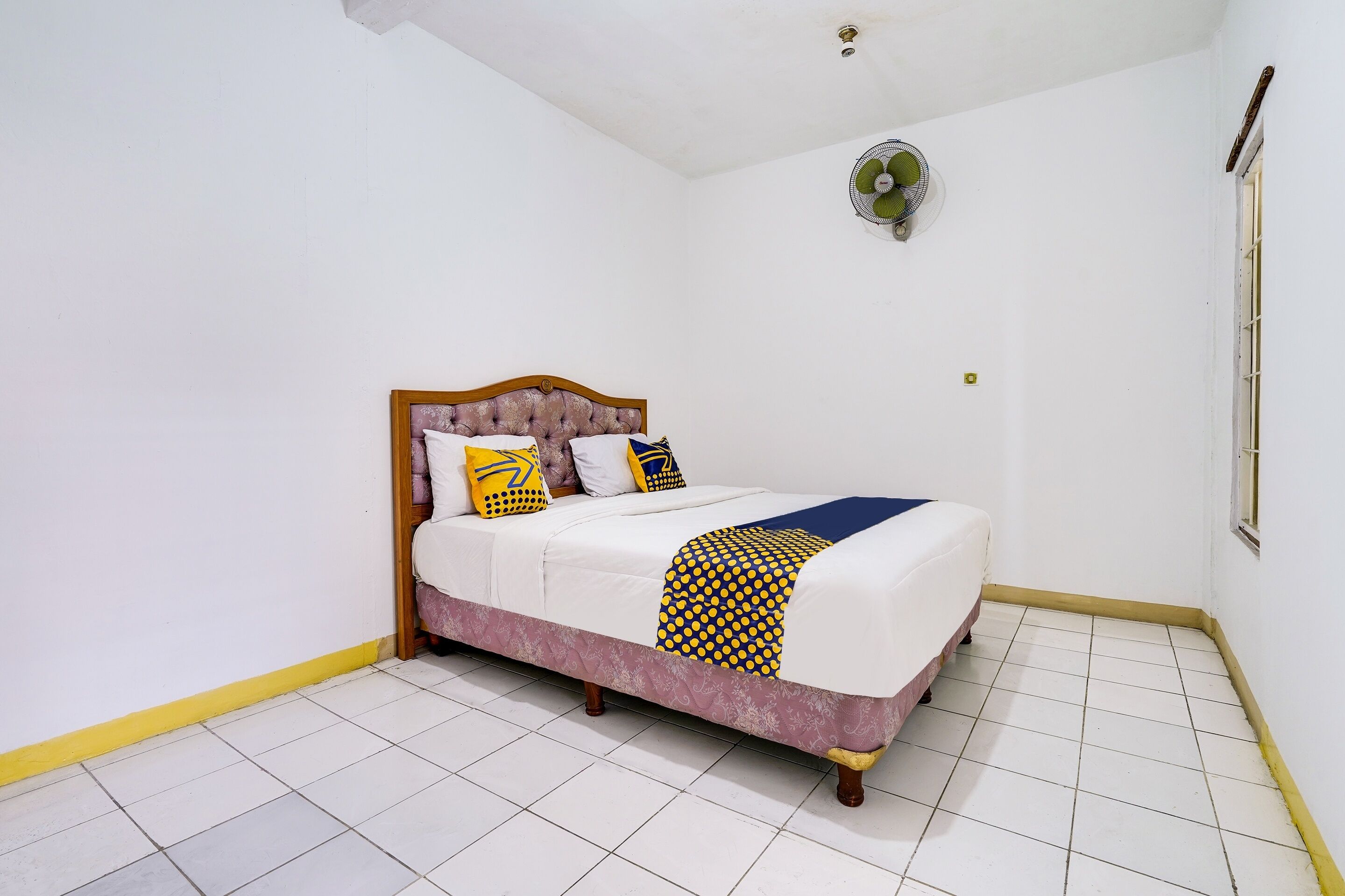 A neatly arranged bedroom with a bed, a wall-mounted fan, and a window with bars.