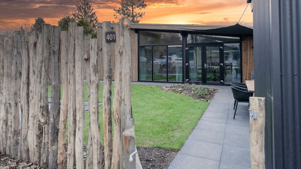 A modern building with a glass entrance, a wooden fence with a sign reading "401", and a paved pathway leading to a seating area. - Child-friendly house with trampoline (Voorthuizen)