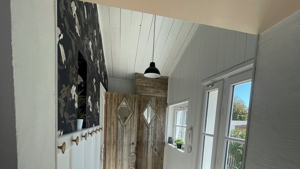 A hallway with white walls, a wooden door, and a window. There are two wooden stools and a hanging light fixture.