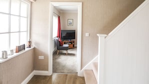 A hallway with a carpeted floor, a window with a curtain, and a door leading to a living room with a TV and a red chair. - Garth (Pwllheli)