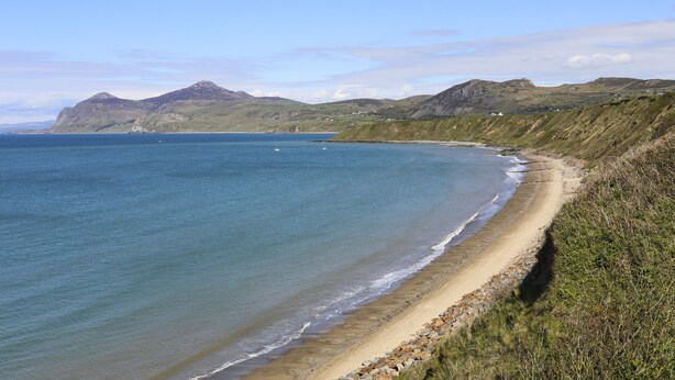 A coastal landscape with a sandy beach, clear blue sea, and distant mountains. - Garth (Pwllheli)