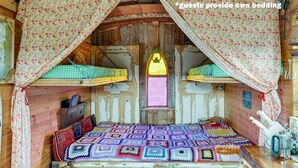 Une cabane en bois rustique, avec un lit recouvert d’une couverture colorée, une lanterne suspendue au plafond et un panneau indiquant : « les clients doivent apporter leur propre literie ».