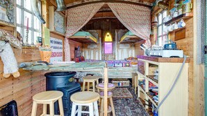 L’intérieur d’un chalet rustique comprend un coin repas, des tabourets en bois, un poêle et un comptoir de cuisine où sont disposés divers objets.