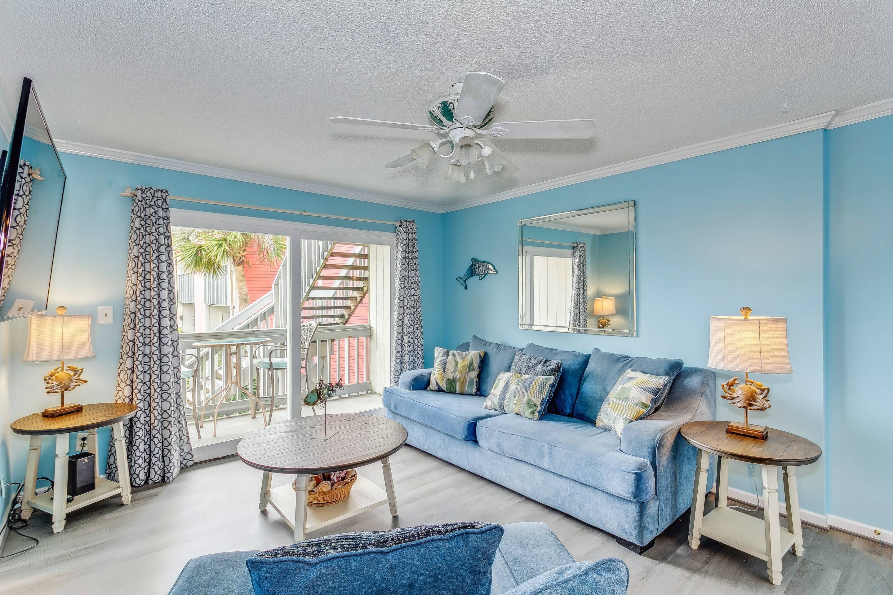 A living room with a blue sofa, two side tables with lamps, a ceiling fan, and a view of a balcony with curtains.