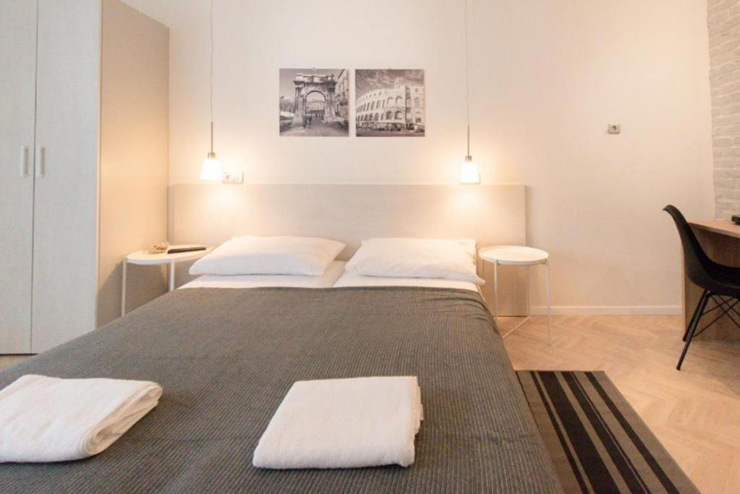 A modern hotel room with a large bed, two bedside tables, a desk, and a chair. There are two wall-mounted lights and two framed pictures above the bed.