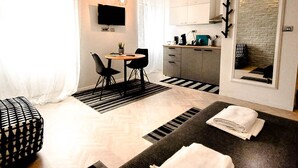 Habitación de hotel compacta con cama, una pequeña mesa de comedor, una silla, un sofá, televisión, área de cocina y un espejo grande.