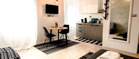 A compact hotel room with a bed, a small dining table, a chair, a sofa, a TV, a kitchen area, and a large mirror.
