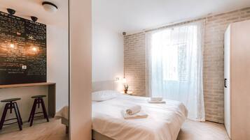 Une chambre d’hôtel moderne avec un lit, un bureau avec des tabourets, un mur en briques et une grande fenêtre ornée de rideaux légers.