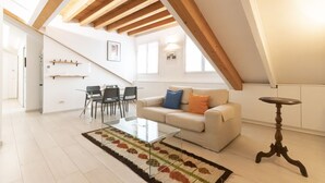 Ein geräumiger Loft mit hoher Decke, freiliegenden Holzbalken, einer Couch mit Kissen, einem Glastisch und einem Essbereich mit Stühlen.