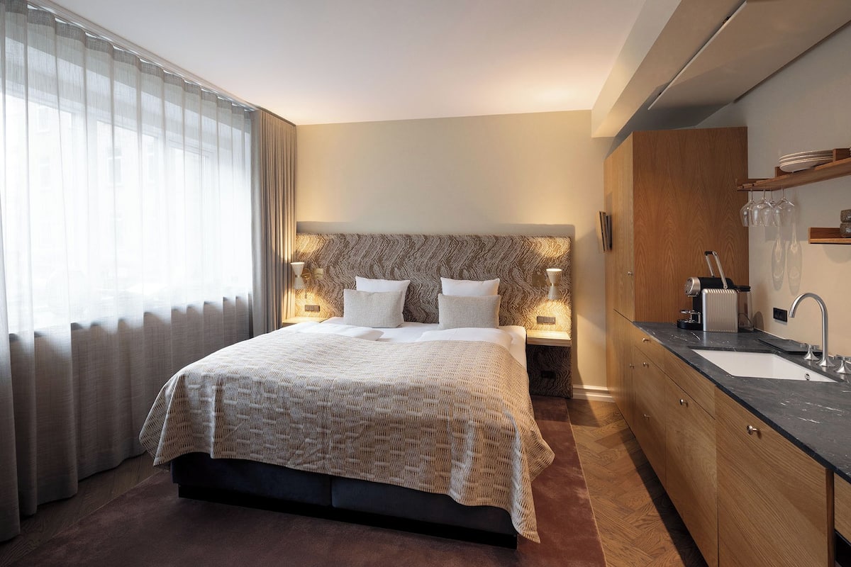 Ein Hotelzimmer mit Bett, Nachttischlampen, einem Holz-Kopfteil, einem großen Spiegel, einem Waschbecken und einem Fenster mit Vorhängen.