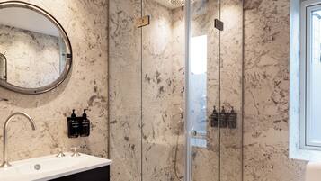Un baño moderno con ducha envuelta en vidrio, pared con azulejos de mármol, espejo redondo y lavamanos blanco con dispensadores de jabón negros.