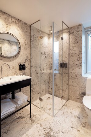 Un bany modern amb dutxa de vidre, paret revestida de marbre, mirall rodó i un lavabo blanc amb dosificadors de sabó negres.