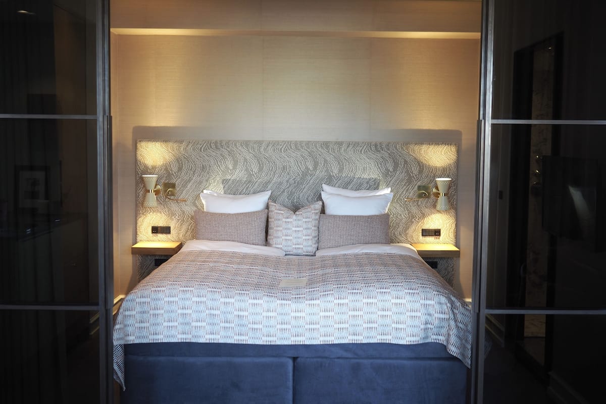 Un dormitorio moderno con un cabecero estampado, una cama grande con muchas almohadas, lámparas de noche y un banco de color oscuro.