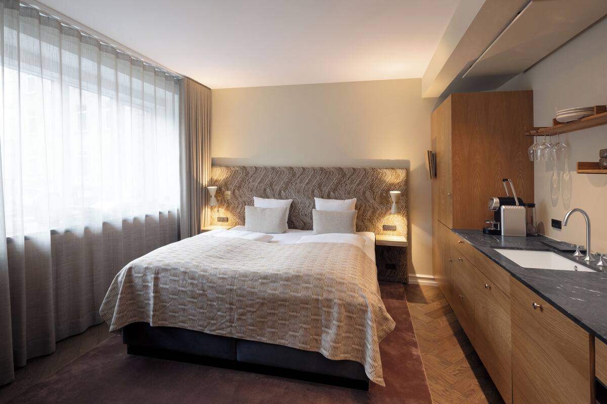 Una habitación de hotel moderna con una cama grande, lámparas de noche, un estante empotrado con vasos y un baño con lavamanos y espejo.
