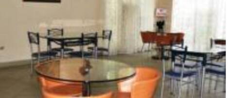 Un comedor con una mesa redonda y sillas naranjas, una mesita con mantel rojo y una ventana con cortinas.
