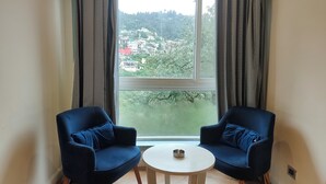 A room with two blue chairs, a small white table, and a large window offering a view of a hillside with buildings. - Hotel Sapling Mussoorie - 500 Mtr from Picture Palace Mall Road (Dehradun)