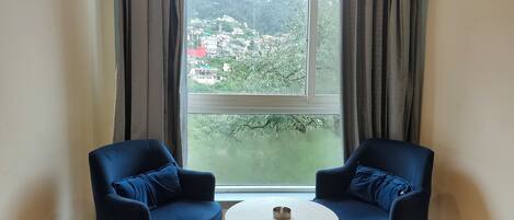 A room with two blue chairs, a small white table, and a large window offering a view of a hillside with buildings.