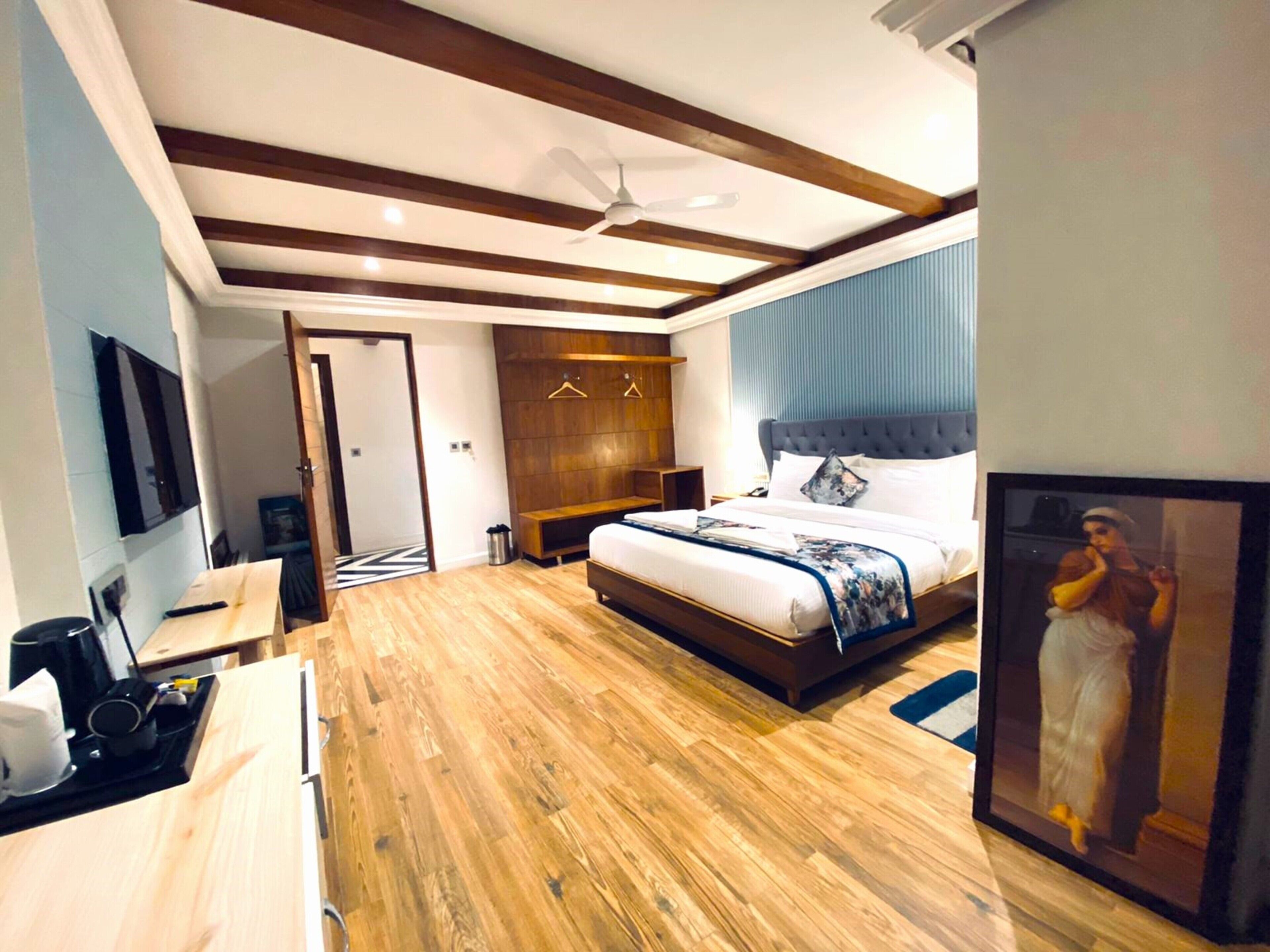 A modern hotel room with a wooden floor, a large bed with a grey headboard, a flat-screen TV mounted on the wall, and a framed picture on the wall.