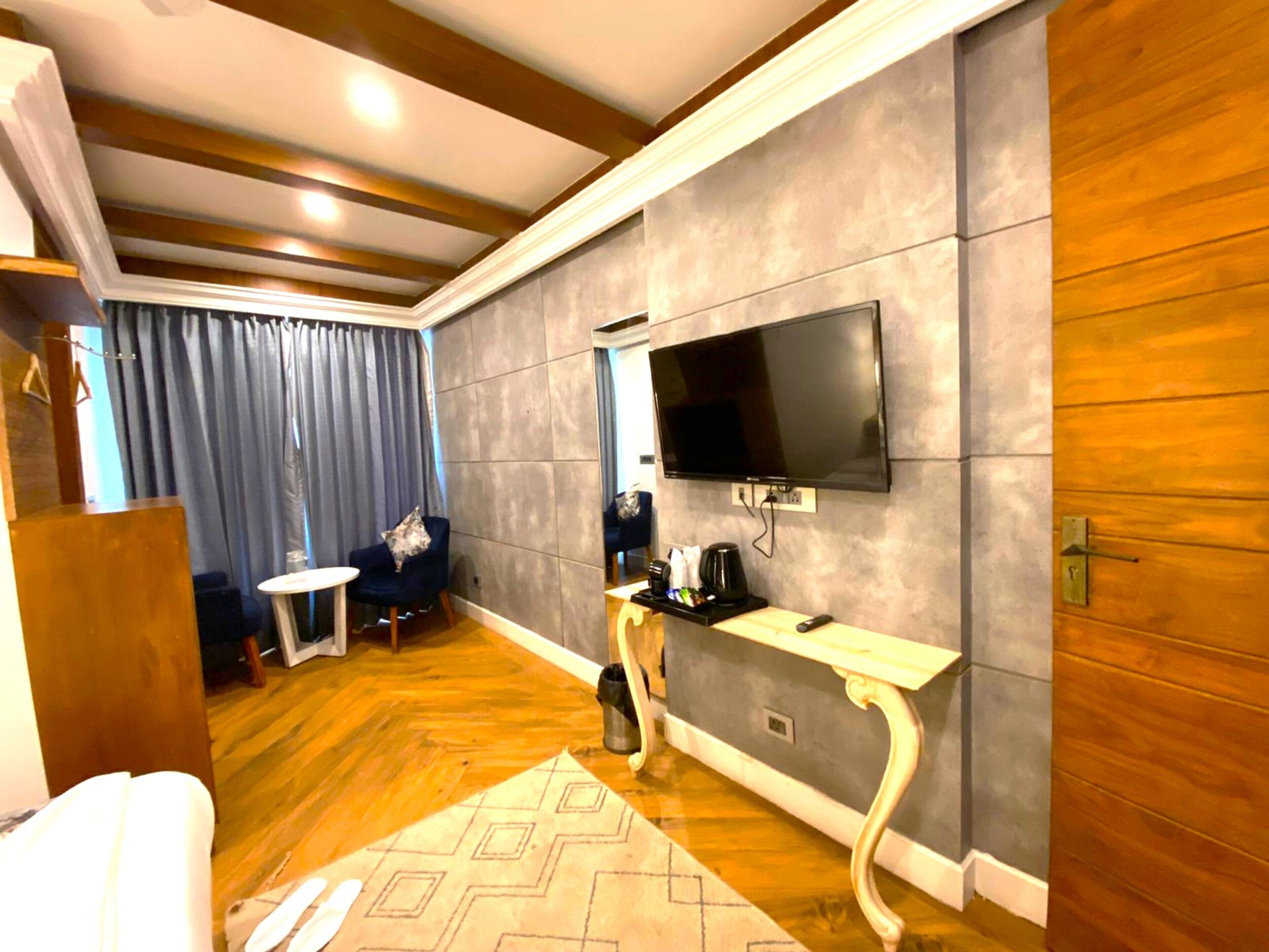 A hotel room with a wooden floor, a grey textured wall, a flat-screen TV mounted above a small table, a sofa, and a coffee machine.