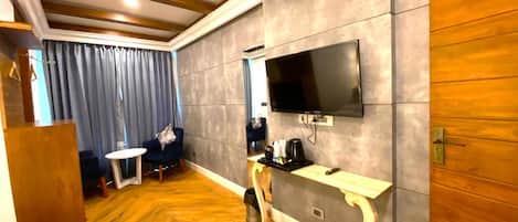 A hotel room with a wooden floor, a grey textured wall, a flat-screen TV mounted above a small table, a sofa, and a coffee machine.