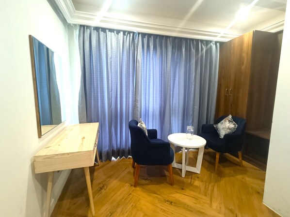 A room with a wooden floor, a desk, two chairs, a small round table, and a large window with blue curtains. - Hotel Sapling Mussoorie - 500 Mtr from Picture Palace Mall Road (Dehradun)