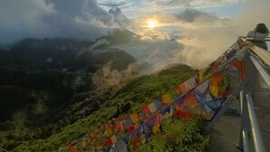 A mountainous landscape with a sunrise, a railing with colorful prayer flags, and a person standing on the railing. - Hotel Sapling Mussoorie - 500 Mtr from Picture Palace Mall Road (Dehradun)