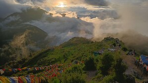 A mountainous landscape with a clear sky, clouds, and a sun peeking through. There are colorful prayer flags and a few people in the distance. - Hotel Sapling Mussoorie - 500 Mtr from Picture Palace Mall Road (Dehradun)