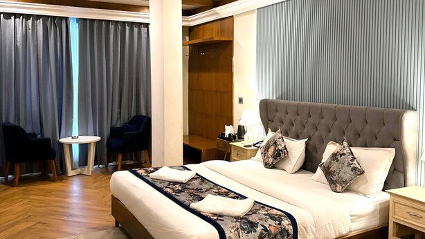 A spacious hotel room with a large bed, a ceiling fan, a wooden floor, and a view of the outdoors through the curtains. - Hotel Sapling Mussoorie - 500 Mtr from Picture Palace Mall Road (Dehradun)