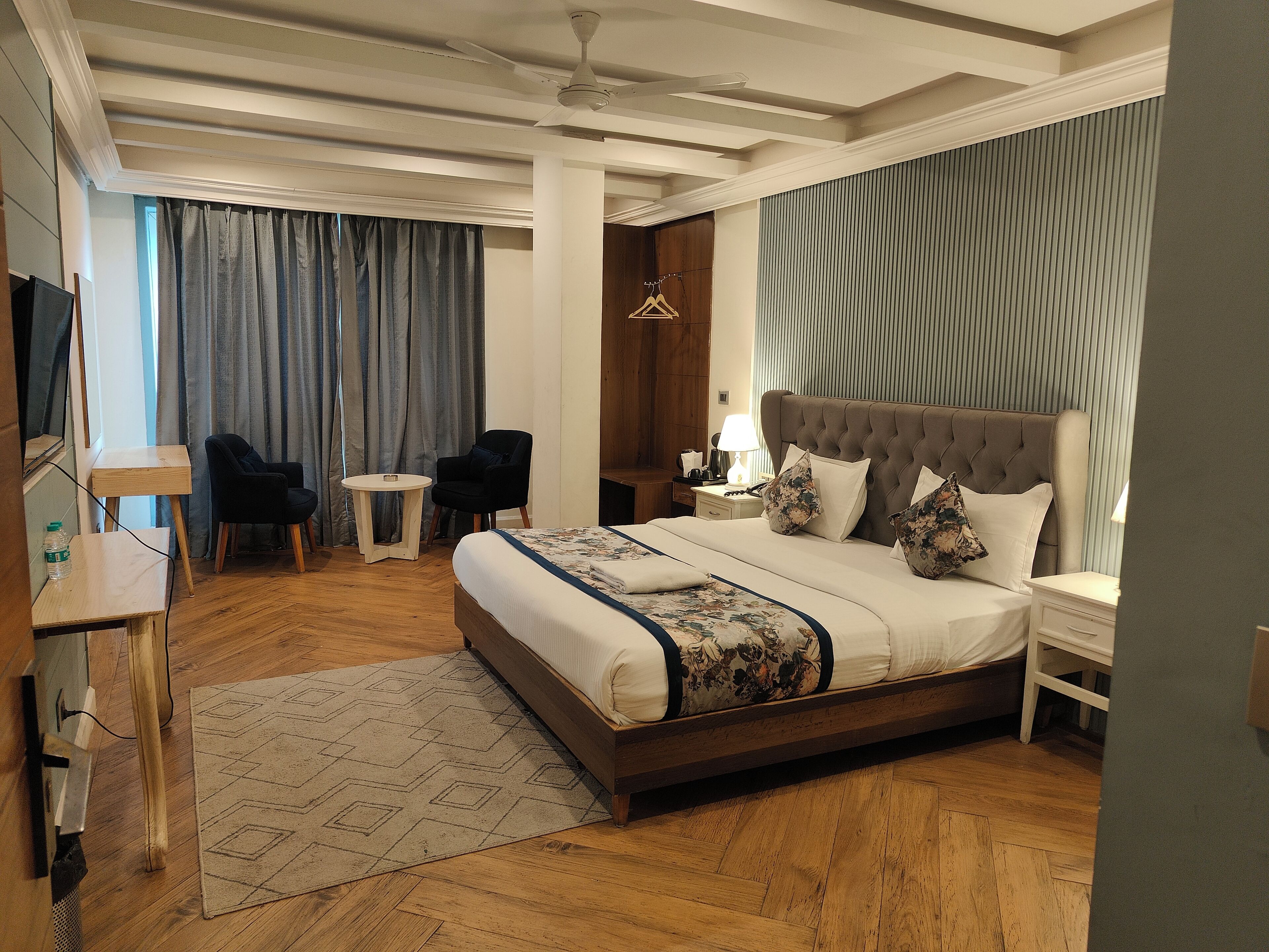 A hotel room with a large bed, a small round table, a chair, a TV, and a desk with a lamp.