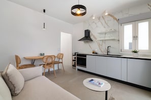 Une cuisine moderne avec des armoires blanches, une hotte noire et un dosseret en marbre. On y trouve un coin repas avec une table et des chaises en bois, ainsi qu’un canapé avec un coussin.