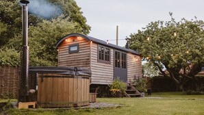 A wooden cabin with a hot tub and a chimney emitting smoke, surrounded by lush greenery.