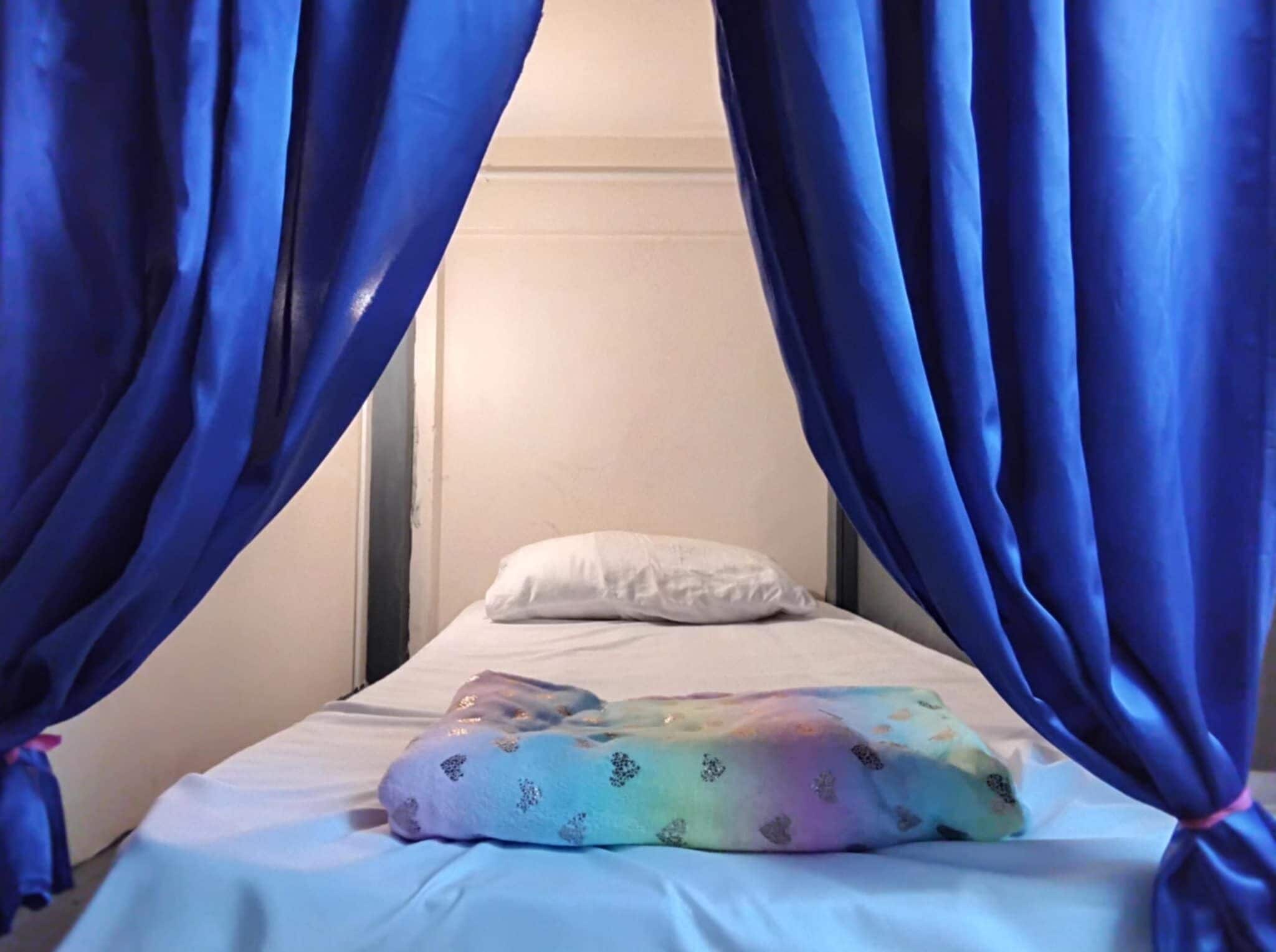 Una cama con un almohadón blanco y una manta de colores del arcoíris, enmarcada por cortinas azules.