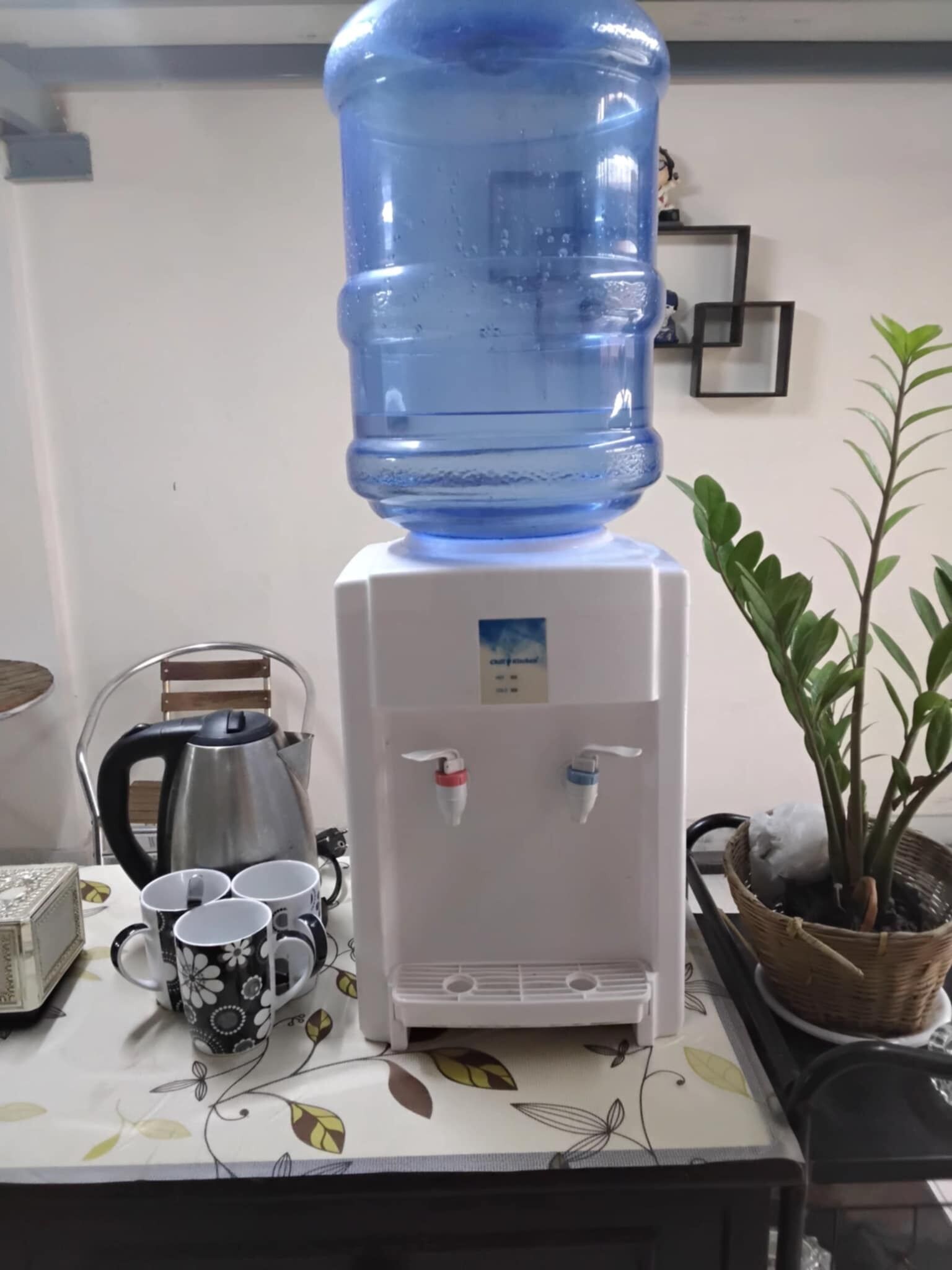 A water dispenser with a large water jug on top, a kettle, and a mug on a table with a floral tablecloth.