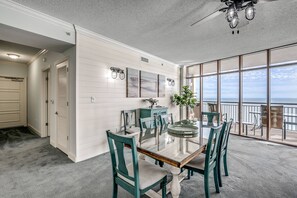 Un espace repas avec une table et des chaises en verre, un mur blanc orné d’œuvres d’art et une vue sur l’océan grâce à de grandes fenêtres.