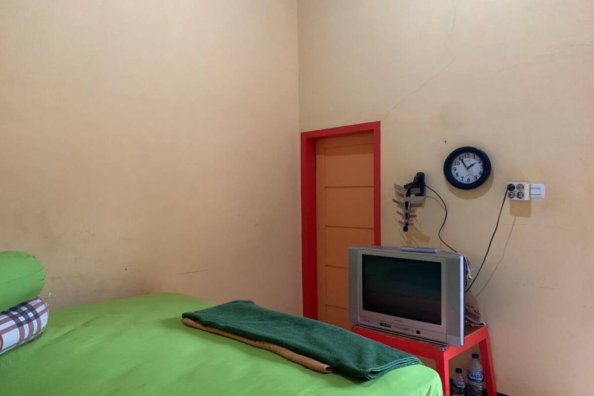 Una habitación con una cama, un televisor sobre un mueble bajo, un reloj en la pared y una puerta.