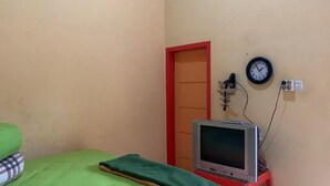 Una habitación pequeña con una cama, un televisor sobre un soporte, un reloj en la pared y una puerta con marco rojo.
