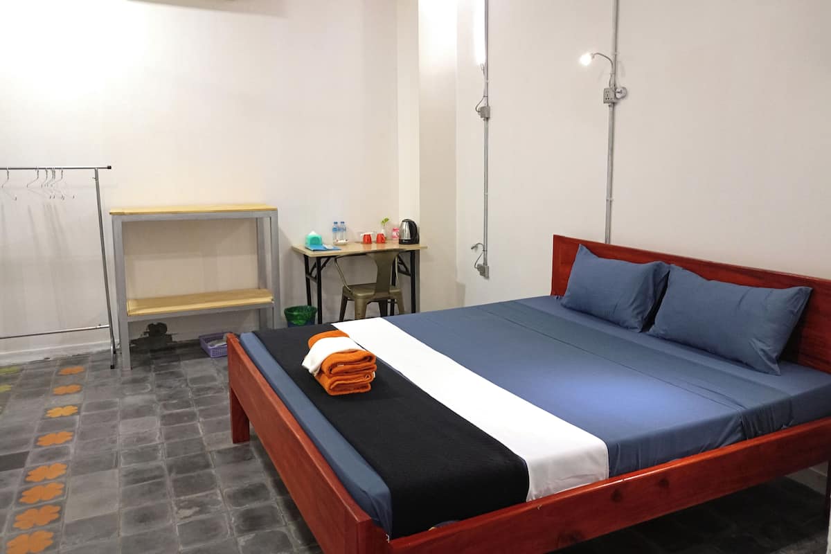 A single-bed room with a wooden frame, a desk with a chair, a small table, and a wall-mounted air conditioner.