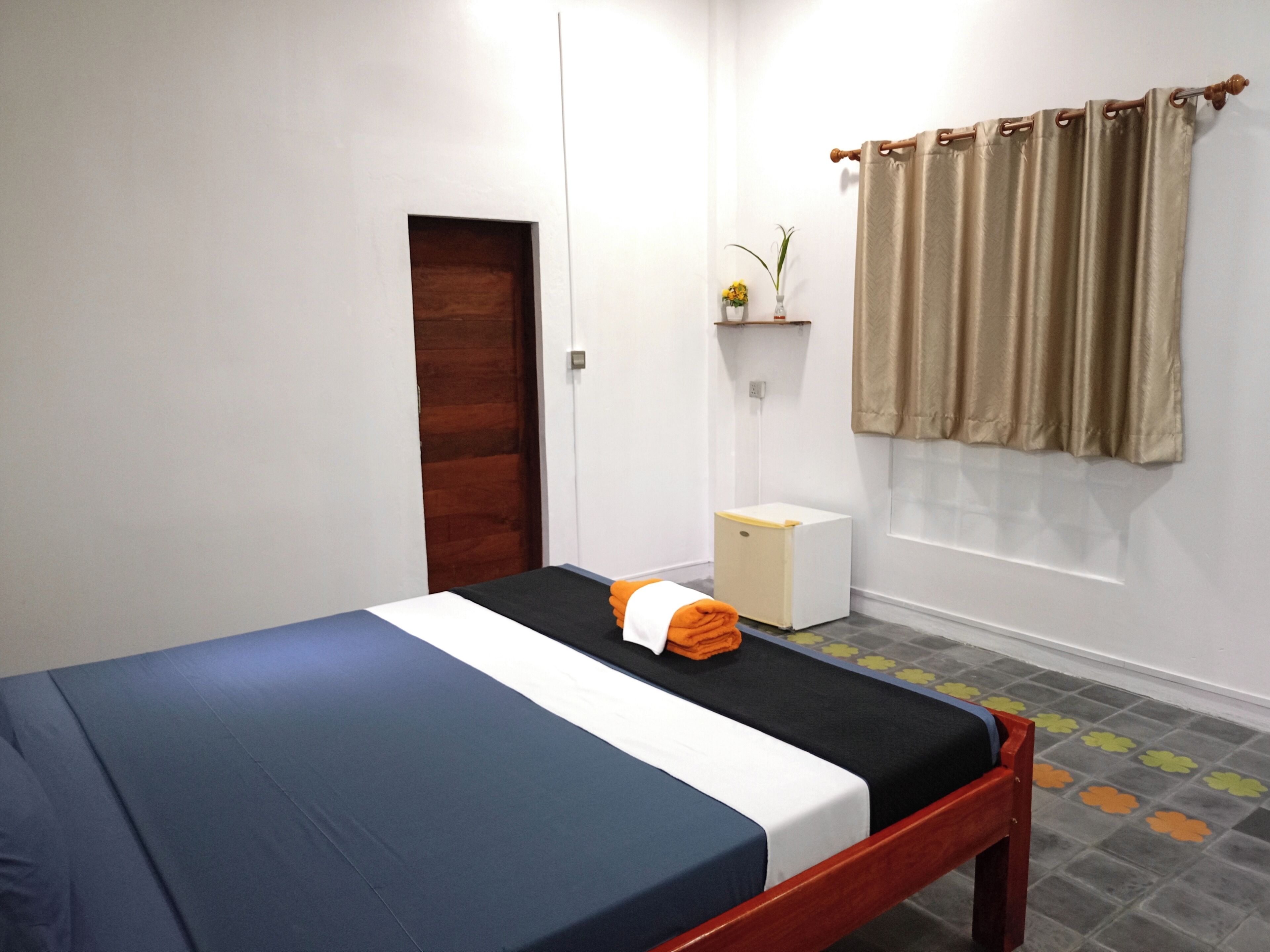 A single-bed room with a wooden frame, a white door, a small white cabinet, a beige curtain, and a flower vase on a shelf.