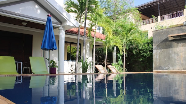 A clear swimming pool with lounge chairs and a blue umbrella, surrounded by palm trees and a building with a red-tiled roof. - Passport Villa (Siem Reap)