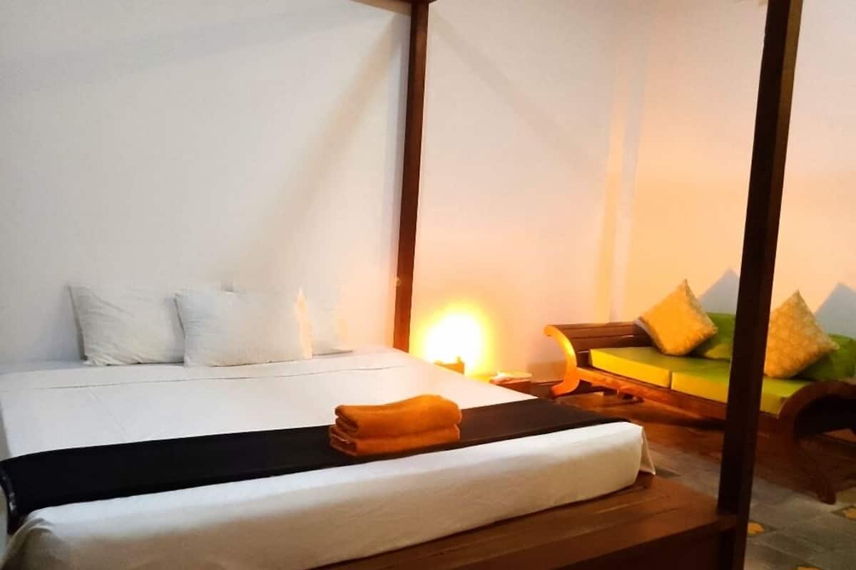 A four-poster bed with white linens, a folded orange towel, and a bedside table with a lamp.