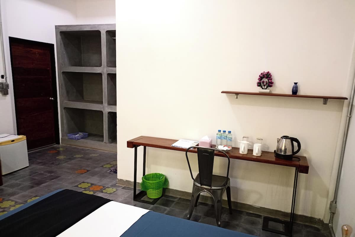 A room with a bed, a desk with a chair, a shelf with a vase, and a trash bin.
