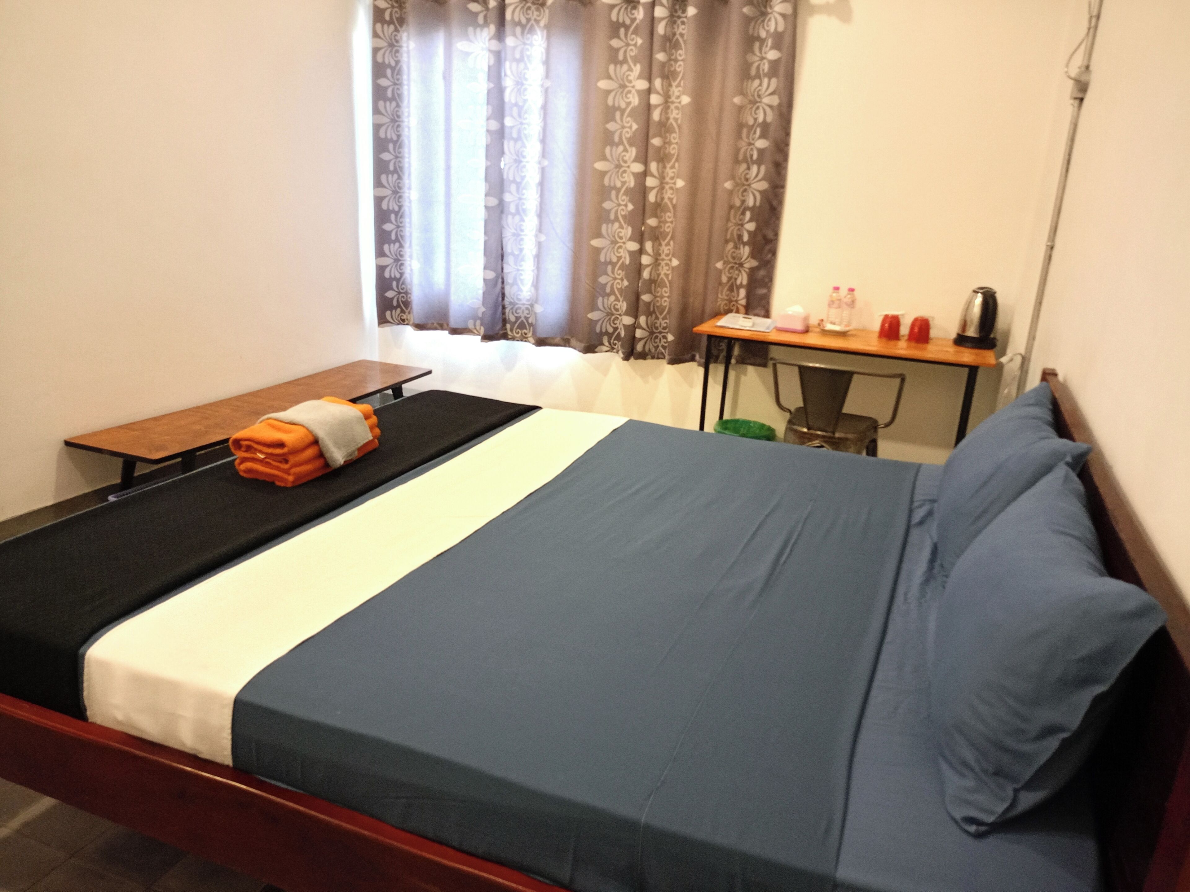 Un letto singolo con copriletto blu e bianco, una panca di legno con asciugamani, una scrivania con sedia e una finestra con tende floreali.