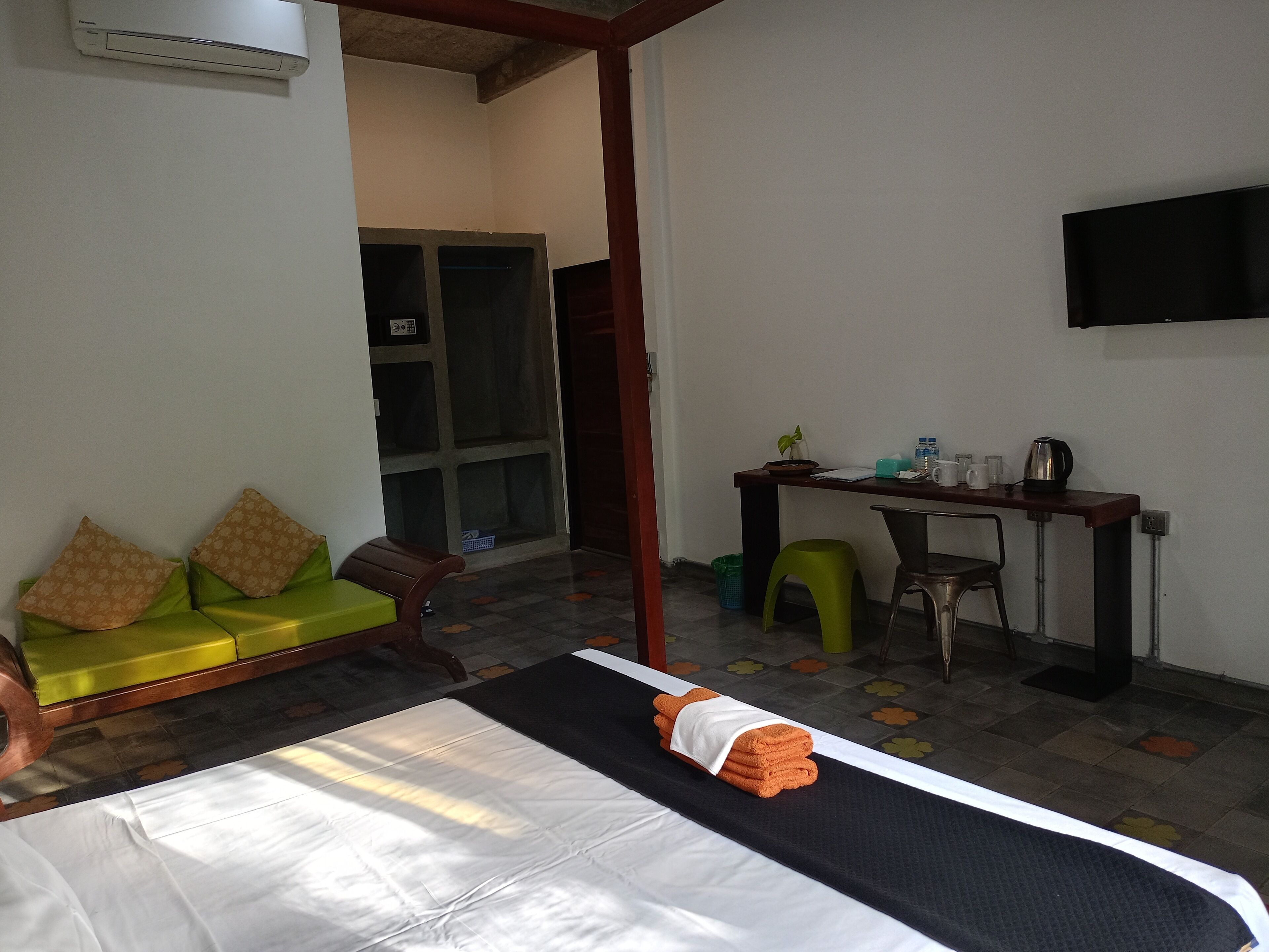 A hotel room with a bed, a green sofa, a wooden chair, a desk with a chair, a television on the wall, and a small plant on the desk.