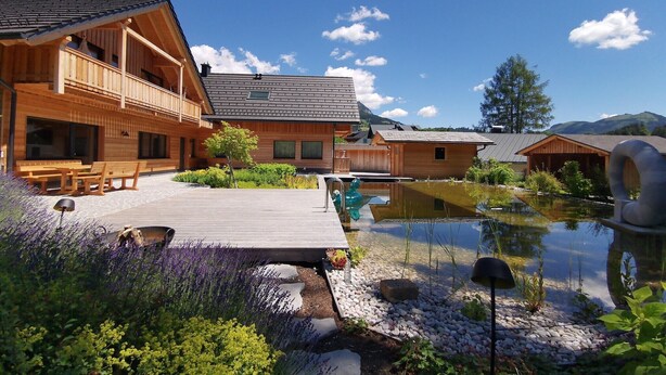 A wooden cabin with a deck, a pond, and surrounding greenery.