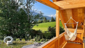A wooden deck with a hammock, a circular seating area, and a view of a green field and mountains.