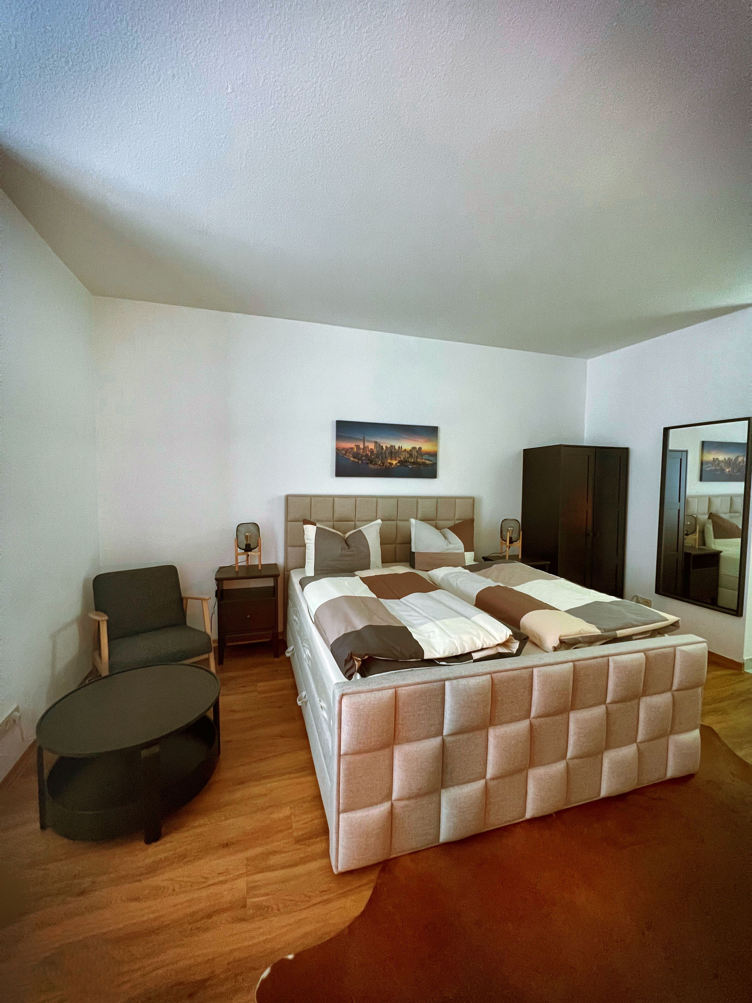 Quarto com uma cama grande, uma cadeira, uma mesa pequena, um espelho e um quadro na parede.
