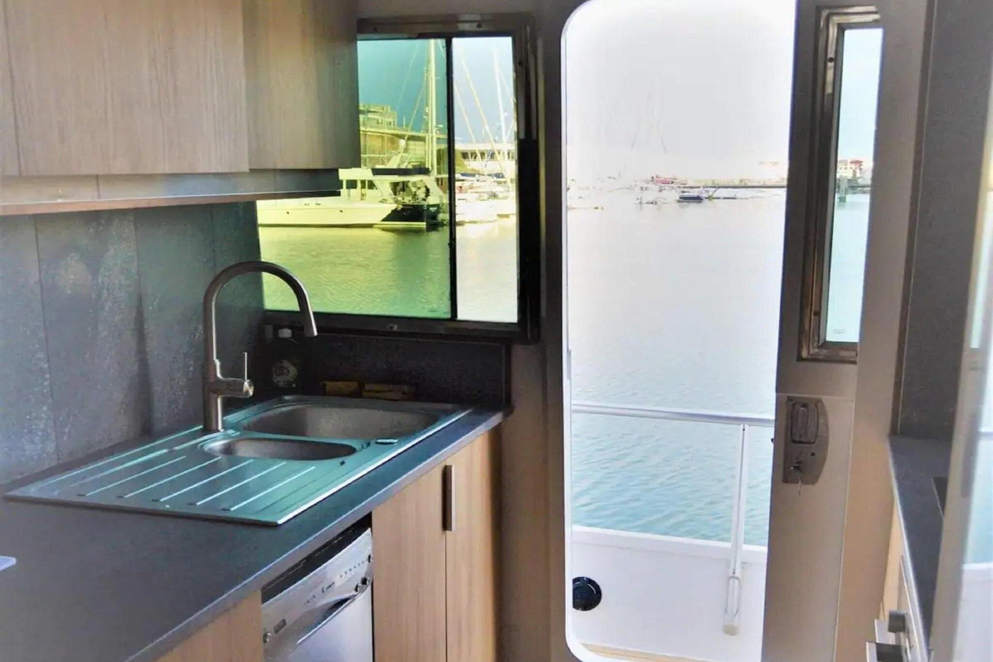 Une petite cuisine équipée comprenant un évier, une fenêtre donnant sur un port de plaisance et une vue sur les bateaux et les bâtiments.